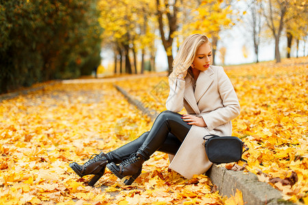 穿着时髦外套穿着时髦鞋子和手提包的漂亮时尚年轻金发女人坐在公园里图片