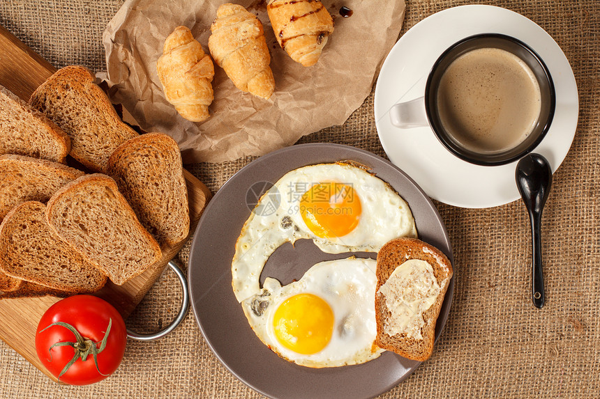 带黄油煎蛋吐司一杯黑咖啡羊角面包木制切板上的面包新鲜番茄的盘图片