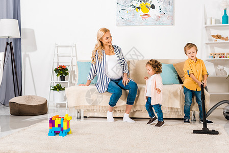 微笑着的孕妇看着可爱的小孩在家中用吸尘器清图片