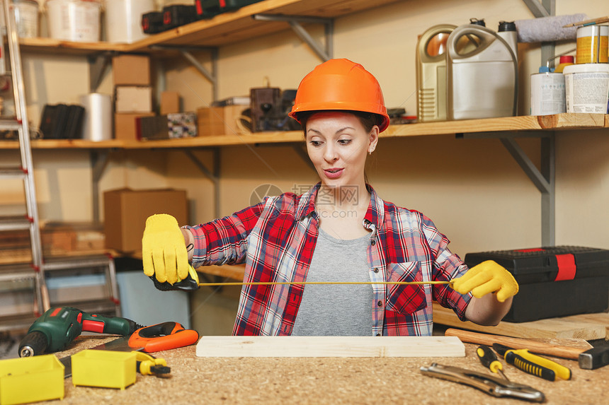 穿着格子衬衫灰色衬衫黄色手套用卷尺测量木头长度的防护头盔在木桌地点的木工车间用不同工具图片