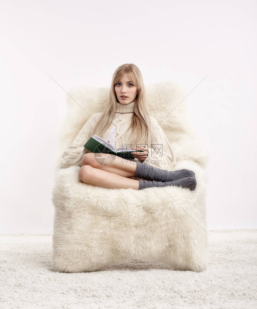 手里拿着书的金发美女的画像女穿着白色羊绒衫和羊毛袜女孩坐在白色毛皮扶手椅上图片