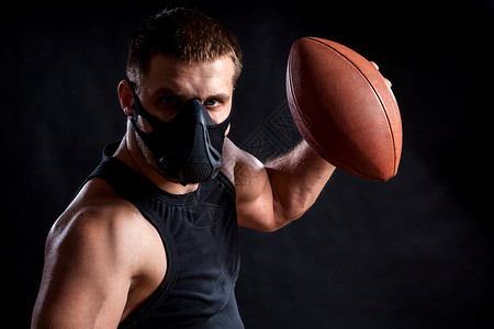 青年运动男子身穿运动衬衫训练面具和以黑色孤立背景持有橄榄球的图片