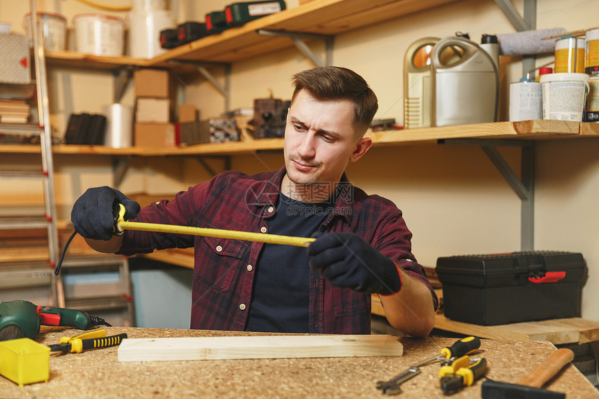 身穿格子衬衫黑色T恤用卷尺测量木头长度的手套在木桌地点的木工车间用不同的工作具作的英俊严肃的图片