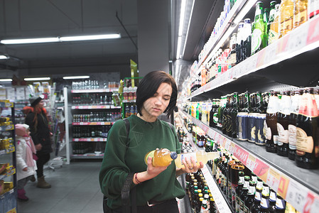 女孩看着她手中的啤酒瓶一个女孩在超市选择酒精饮料商店图片