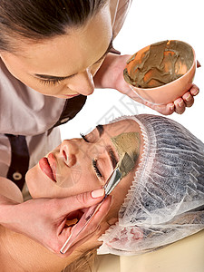 水疗沙龙中女人的泥面膜用粘土全脸按摩与治疗室的女孩美容师与碗治疗程图片