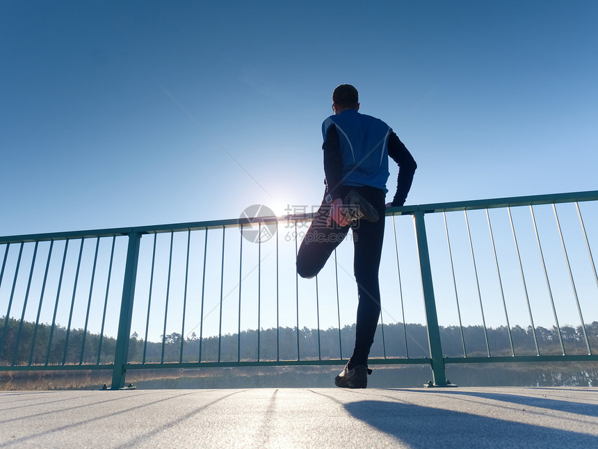 穿着高大黑色紧身裤的跑步者使身体在桥路上伸展太阳正在勾勒人的身体在湖桥上光滑的混凝土地面上图片
