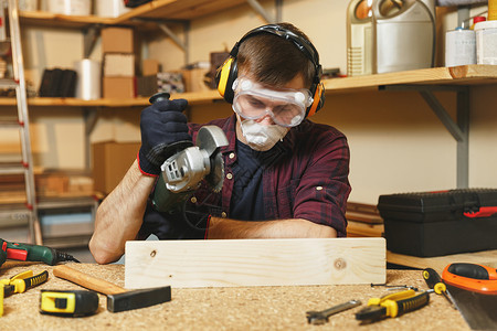 身穿格子衬衫黑色T恤隔音耳机在木桌地点的木工车间用不同工具作的防护面罩用电锯木头背景图片