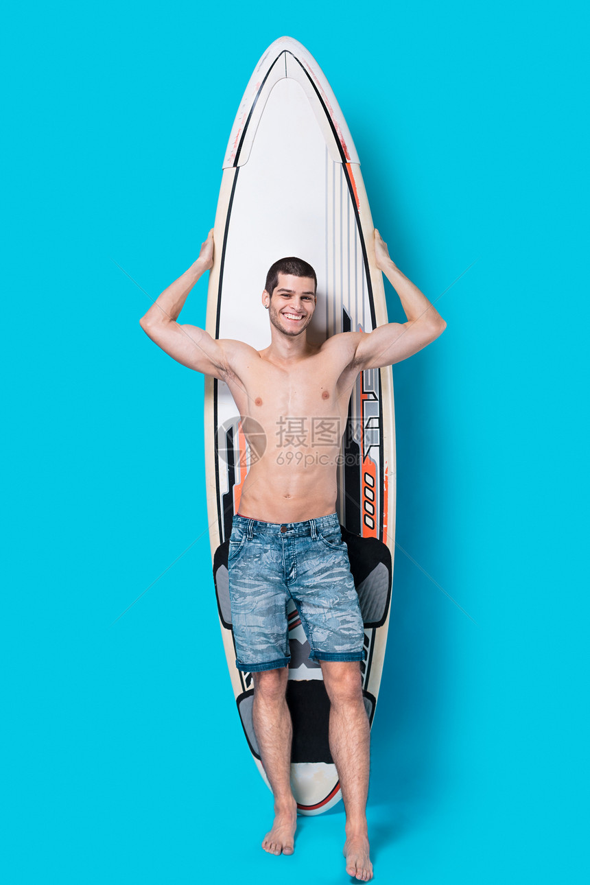 格开朗的人在工作室里与冲浪合影在工作室的蓝色背景上有冲浪板的冲浪者的全长肖像男拿着冲浪板显示图片