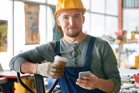 留着胡子的年轻工人在工厂车间休息时用智能手机拿着咖啡杯图片