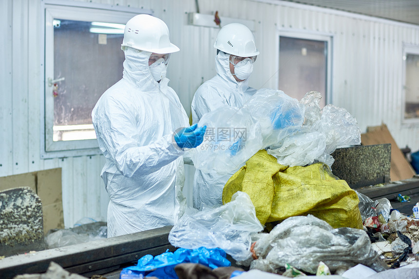 废物处理厂两名身穿生物危害防护服的工人在传送带上分拣可回收塑料图片