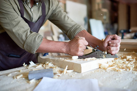 在木匠车间制造家具时用平面工具刮削木头的强壮男图片