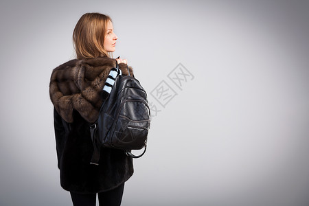 身穿天然貂皮黑色皮大衣的黑发年轻女子手里拿着一个黑色背包图片