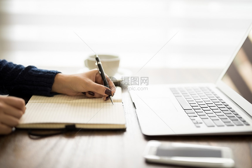 用笔记本电脑在她的日记上写笔记的女人特写图片