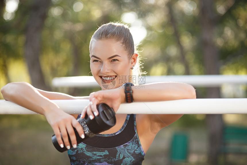 有热水瓶的运动女人英俊的积极微笑的女人与健身锻炼后休息的肌肉发达的身体运动型女运动员坐在公园里图片
