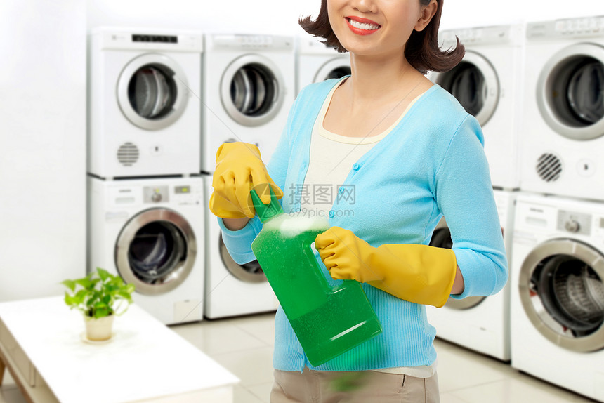 穿着橡皮手套微笑的年轻女在站宽敞自助洗衣间时打开瓶液体洗涤图片