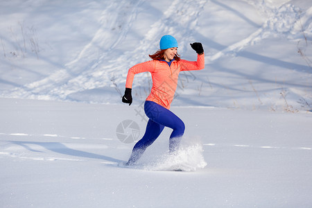 穿着亮蓝帽子橙色衫和裙的年轻女子在寒冬日穿过冬季雪图片