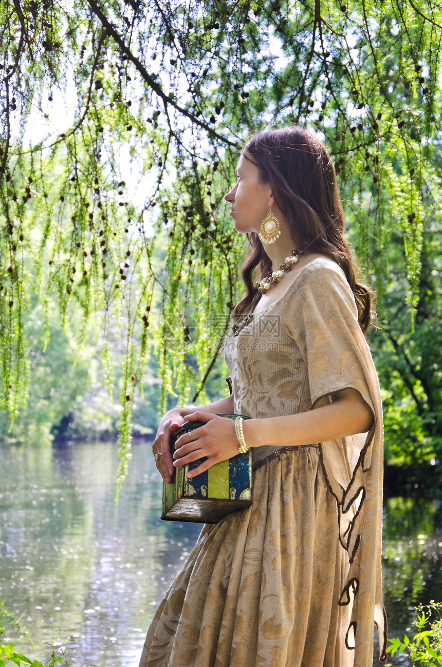 身着旧裙子的年轻女子在公园池塘岸上走来去图片