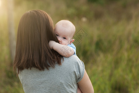 年轻温柔的黑发女人站在绿草背景拥抱可爱的小男孩自然图片