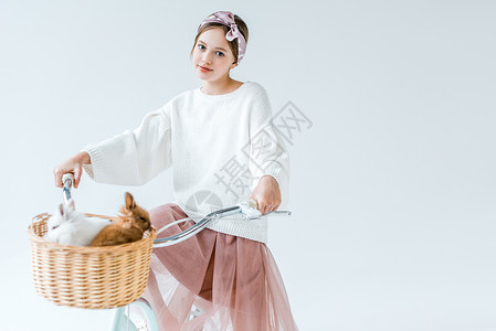 年轻美丽的少女在骑自行车时看着照相机与兔子一起坐在篮子中图片