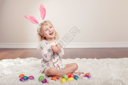 戴着复活节耳朵的可爱有趣的白人小女孩坐在工作室里柔软蓬松的地毯上带着节日彩蛋玩庆祝传统基督背景图片