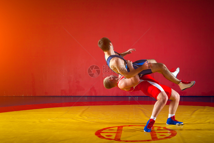 两名身穿蓝色和红色摔跤紧身衣的强壮摔跤手正在健身房的黄色摔跤地毯上摔跤并进行摔跤做图片