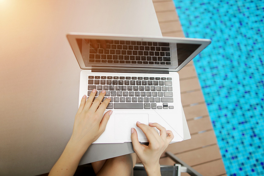 从上面的女人手在阳光明媚的日子游泳池按下笔记本电脑上的按钮的视图图片