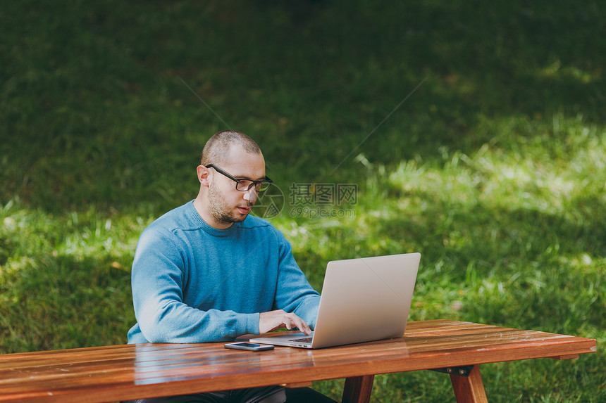 年轻成功的聪明人商或学生在休闲的蓝色衬衫眼镜坐在桌边与手机在城市公园使用笔记本电脑在户外工作在绿色背景图片