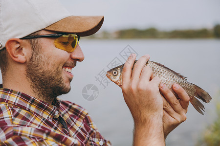 穿着格斗衬衫帽子和太阳镜的年轻无脸微笑的男人在湖边捕到一条鱼图片