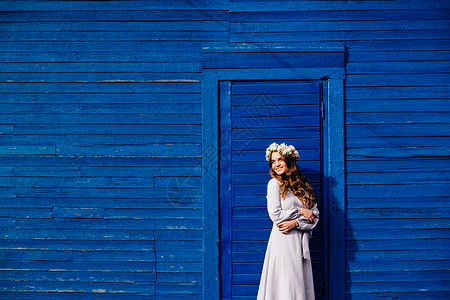 穿着长裙子头上戴花圈的漂亮女孩站在蓝房子图片