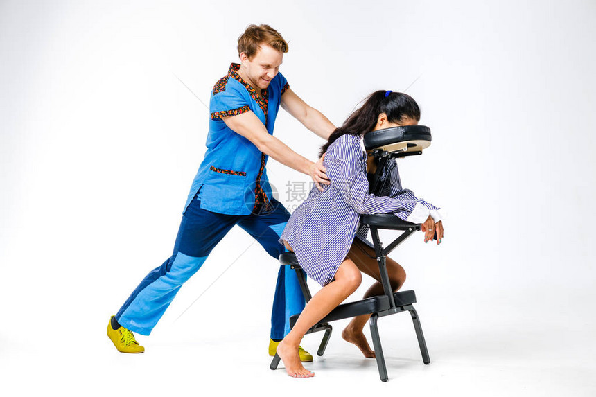 主题按摩和办公室一位身穿蓝色西装面带微笑的治疗师的年轻男子正在为年轻女工穿着衬衫的女商人在指压按摩椅上做背部和颈部按摩图片