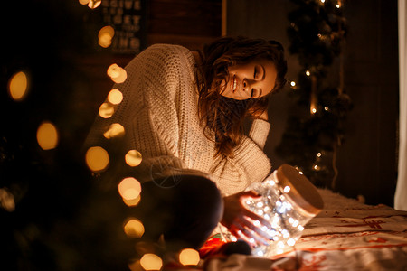 美丽的年轻女孩穿着一件织的长毛衣圣诞前夕在床边装着喜悦灯图片