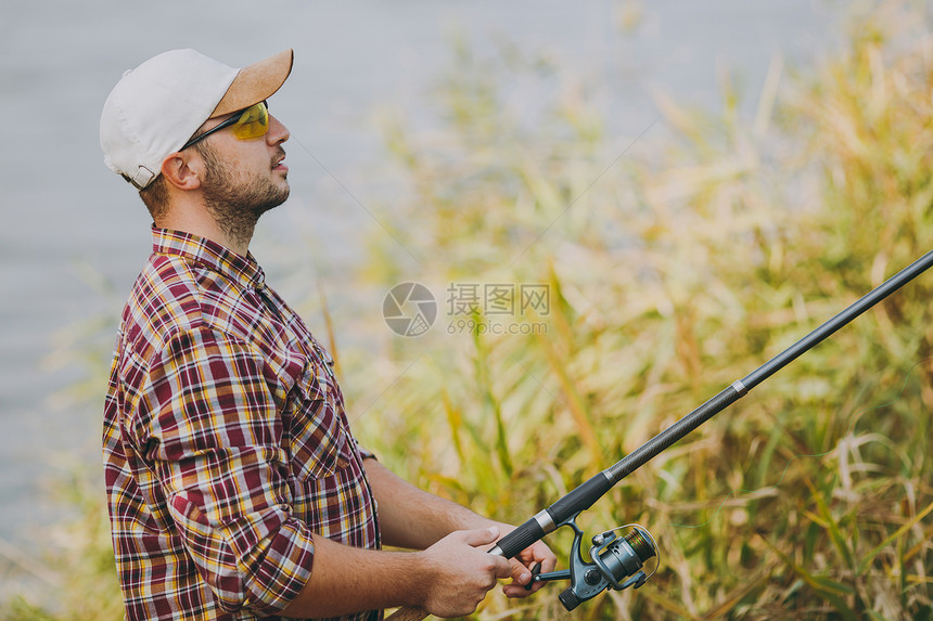 侧视图穿着格子衬衫帽子和太阳镜的年轻未刮胡子的男子在靠近灌木和芦苇的湖岸上拿着一根钓鱼竿和展开的卷轴生活方式休闲图片