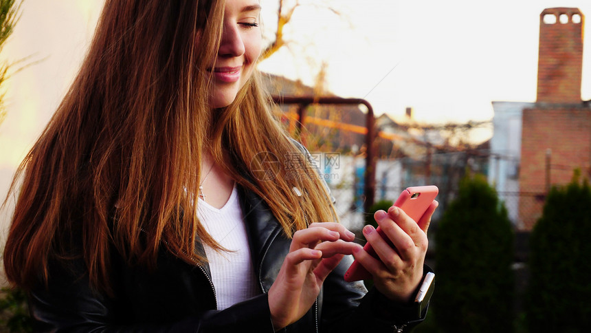 漂亮的女孩在绿色植物附近的粉红色外壳中与智能手机聊天用现代小工具和互联网连接在外面休息的概念美丽的女穿图片