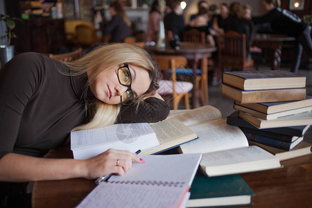 大学的疲倦的年轻女学生在公共图书馆准备考试和学习课程坐在被大量教科书图片