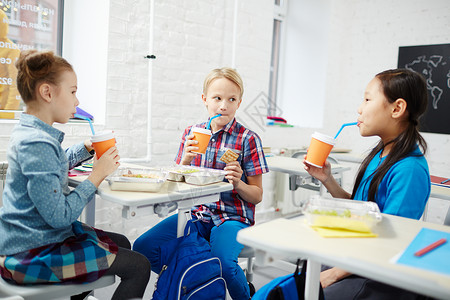 三个小学同在课桌旁吃午饭用塑料杯喝水背景图片