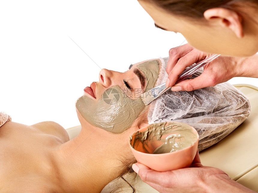 水疗沙龙中女人的泥面膜用粘土全脸按摩与治疗室的女孩美容师与刷治疗程序孤立的背景用于面部的愈图片