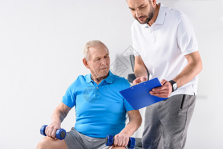 协助老年男子在灰色背景下用哑铃进行锻炼的康复治疗师图片