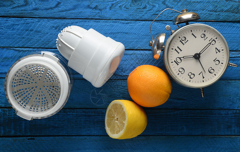 早晨从柠檬和橙子中提取的新鲜果汁手工榨汁机闹钟蓝色木质背景上的柑橘类图片