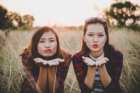 两个漂亮的女孩最好的朋友在夏日落背景的田野上放松愉快地吹嘘着亲吻图片
