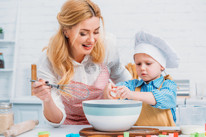 微笑的母亲用手搅拌器和小男孩把鸡蛋倒进碗里图片