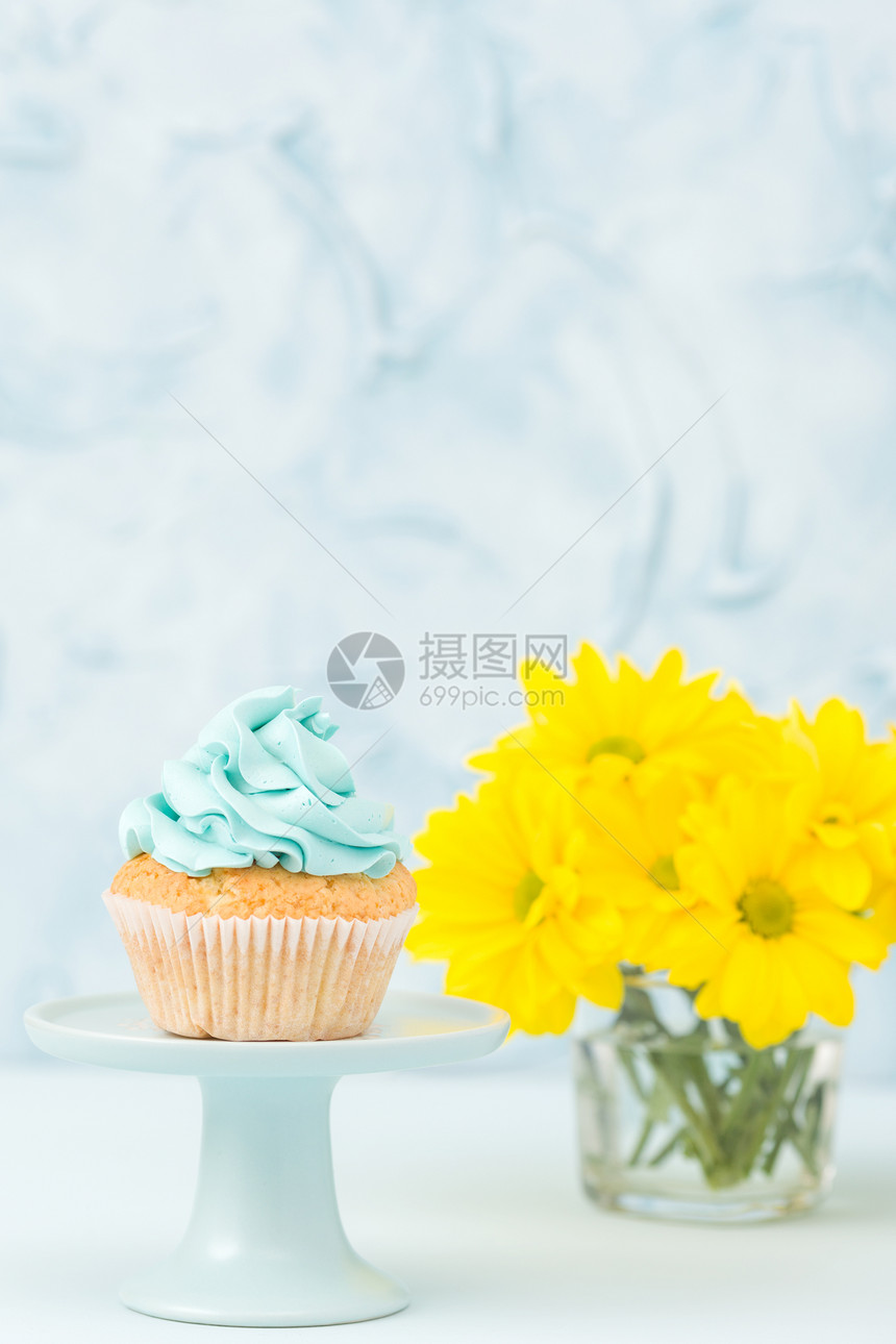 杯形蛋糕与蓝色奶油装饰在立场和黄色菊花束在玻璃花瓶在蓝色柔和的背景与文本区域生日或婚图片