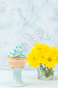 杯形蛋糕与蓝色奶油装饰在立场和黄色菊花束在玻璃花瓶在蓝色柔和的背景与文本区域生日或婚背景图片
