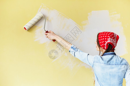 穿着红色头巾的漂亮女孩用油漆滚筒在墙上刷漆一位年轻美女在她的新公寓背景图片