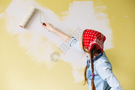 穿着红色头巾的漂亮女孩用油漆滚筒在墙上刷漆一位年轻美女在她的新公寓图片