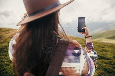 时髦的旅游女郎戴着风毛拿着电话在山顶上拍照图片