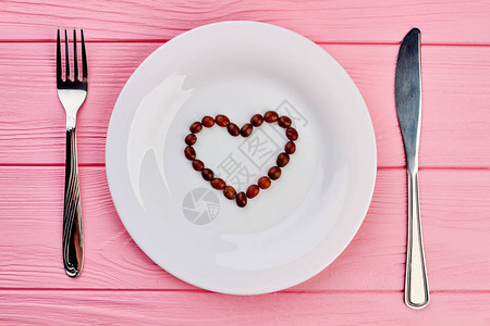 用咖啡豆制成的心白板白色盘子与心形咖啡豆叉子和刀在粉红色的木制图片