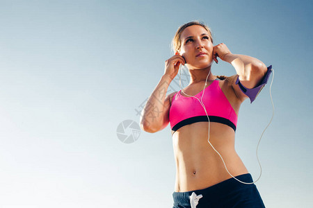使用智能手机操作臂带插件的低角度女运动员在蓝天上放耳机图片