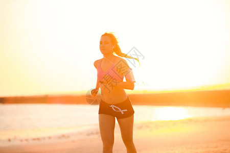 手持耳机智能手机的女运动员在沙滩上与阳光抗日跑步时使用臂背景图片