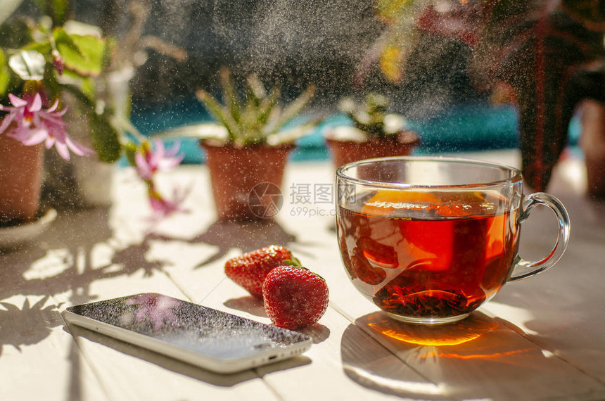 玻璃茶杯红草莓智能手机和在浅木制桌上的锅里放花图片