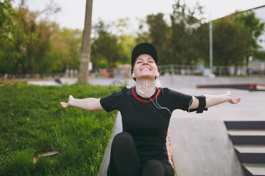 身穿黑色制服带耳机的年轻运动休闲少女听音乐伸出手在跑步前后休息和坐户外城市公园培训图片